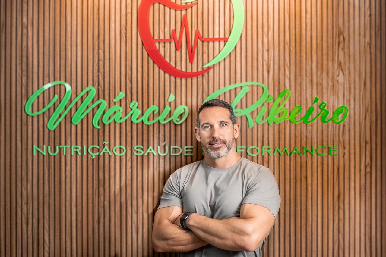 Márcio Ribeiro de braços cruzados, sorrindo, em frente à parede de madeira com a logo verde e vermelha do consultório.